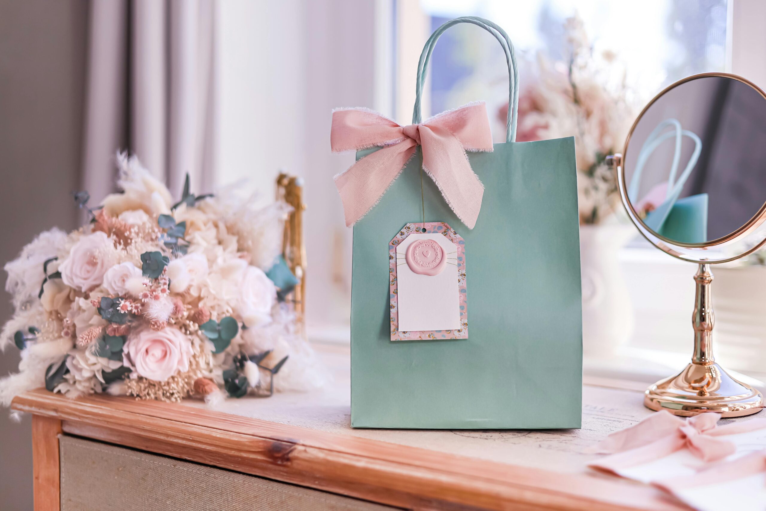 Chic gift bag with ribbon and pastel floral arrangement on a wooden dresser.