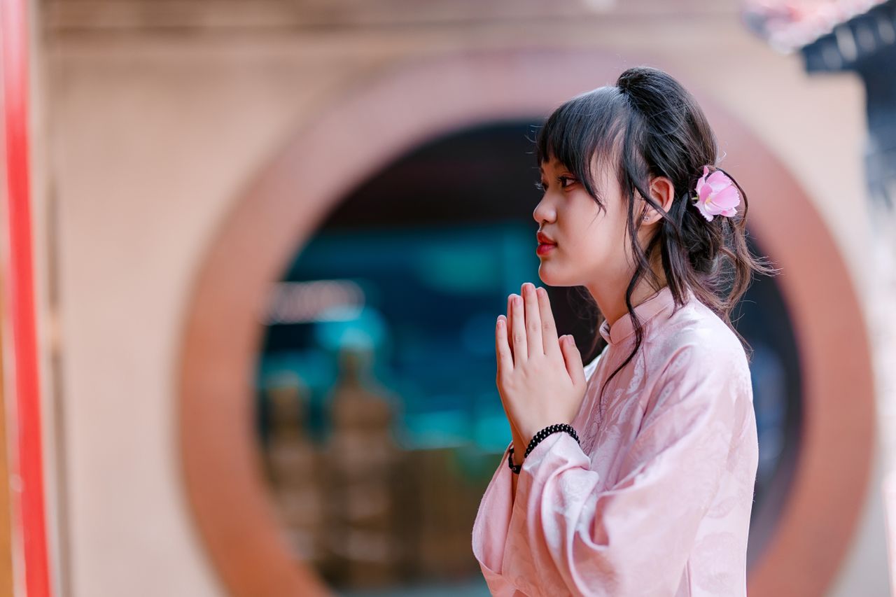 Young Asian woman praying with folded hands in traditional clothing indoors.